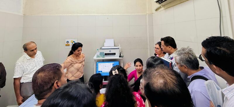 A group of people gathered around a computer workstation as a woman explains something to them in a small office room.