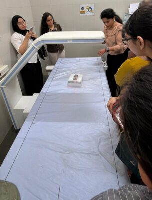 Several women stand around a radiology/medical imaging table in a clinical room, watching a small white box on the table.