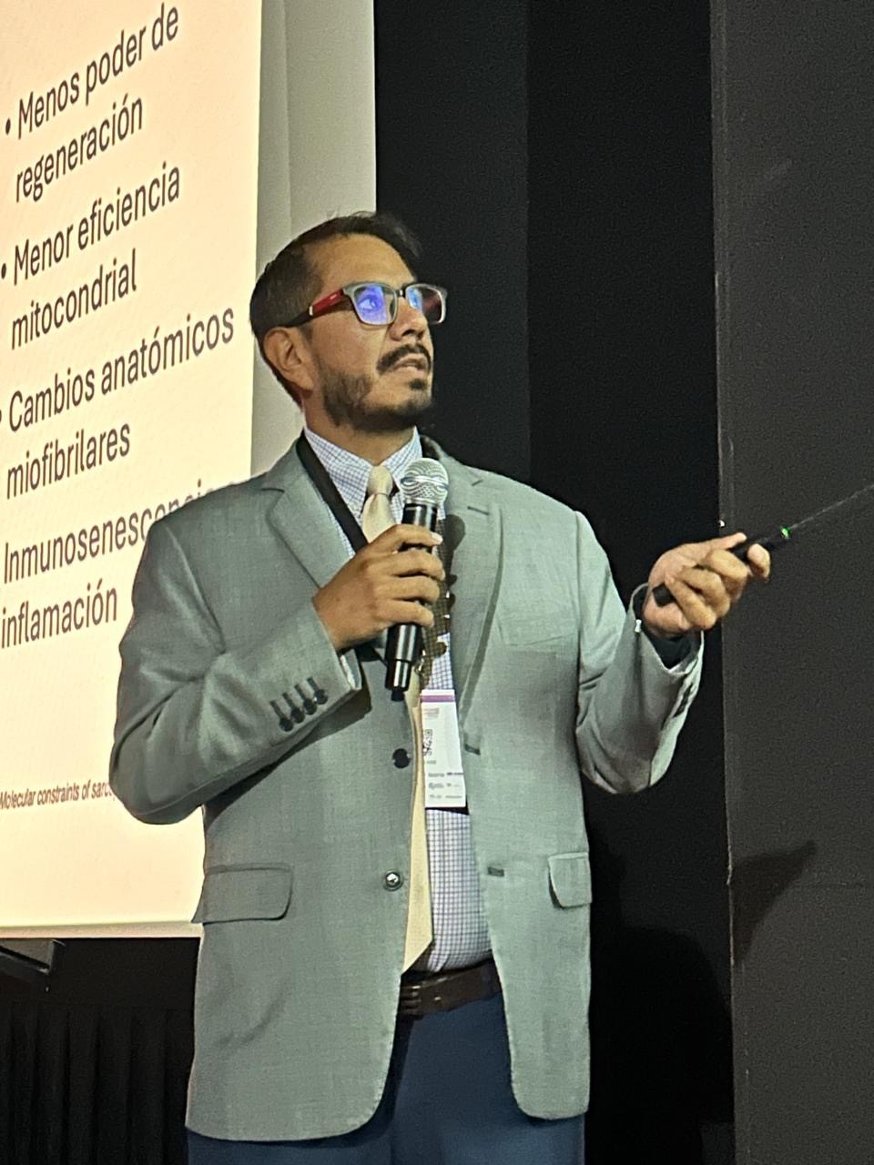 Male presenter in a light gray blazer and blue pants speaks into a handheld microphone on stage, gesturing with a laser pointer as a Spanish slide is projected to the left with bullet points.